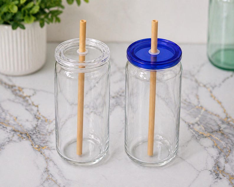 May include: Two clear glass drinking jars with lids and wooden straws. One jar has a clear lid, and the other has a blue lid. The jars are on a marble surface, with a white pot of greenery in the background.