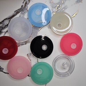 May include: A collection of colorful, round lids in various shades, including red, pink, blue, black, and clear. Each lid has a central, circular design. The lids are arranged on a white and gray marble-patterned surface.