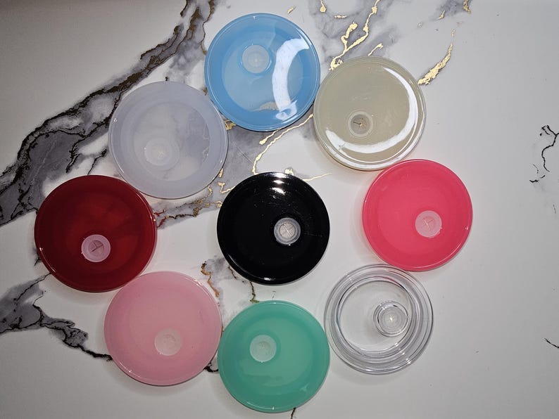 May include: A collection of colorful, round lids in various shades, including red, pink, blue, black, and clear. Each lid has a central, circular design. The lids are arranged on a white and gray marble-patterned surface.