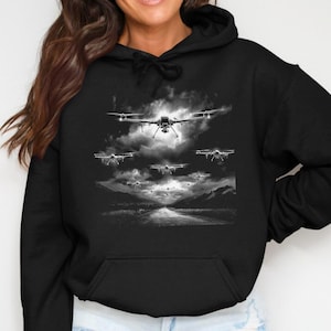 Könnte beinhalten: Schwarzer Kapuzenpullover mit einem Graustufen-Design von Drohnen, die in einem bewölkten Himmel fliegen. Das Design umfasst eine Straße oder einen Wasserweg im Vordergrund und Berge im Hintergrund. Der Kapuzenpullover hat eine Vordertasche und eine Kapuze mit Kordelzug.
