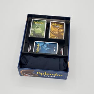 May include: A rectangular board game box for Splendor Duel containing three stacks of playing cards in black plastic inserts and a dark blue fabric drawstring pouch. The box features the game title Splendor Duel in yellow text.
