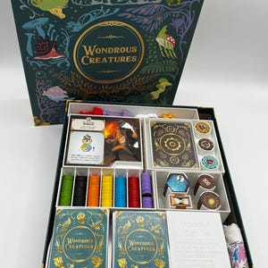 May include: An open board game box with the title "Wondrous Creatures" on the lid and two card decks. The box is teal with gold accents and illustrations of fantastical creatures. Inside are various game components, including cards, tokens, and dice.