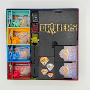 May include: A board game box insert for Drillers containing four coloured plastic trays holding miniature figures and cards, a black foam insert with the word DRILLERS printed in yellow, and black plastic compartments for dice, tokens, and cards.