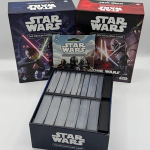 K&ouml;nnte beinhalten: Eine Sammlung von Star Wars Deckbuilding-Spielen. Die Schachteln sind marineblau und rot, mit Bildern von Charakteren, die Lichtschwerter schwingen. Die Spielboxen und Kartenpackungen werden mit einer Kartenbox im Vordergrund pr&auml;sentiert.
