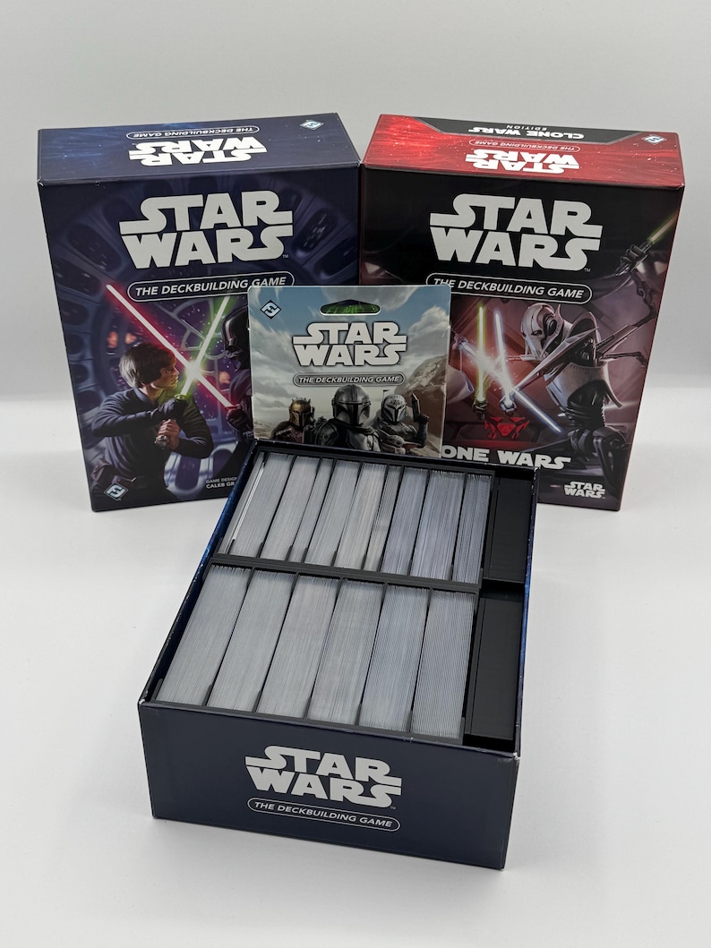 K&ouml;nnte beinhalten: Eine Sammlung von Star Wars Deckbuilding-Spielen. Die Schachteln sind marineblau und rot, mit Bildern von Charakteren, die Lichtschwerter schwingen. Die Spielboxen und Kartenpackungen werden mit einer Kartenbox im Vordergrund pr&auml;sentiert.
