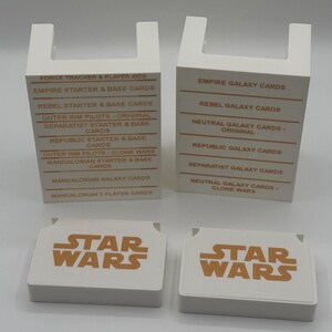 K&ouml;nnte beinhalten: Vier wei&szlig;e Star Wars Kartenboxen und zwei Kartenstapel. Die Boxen haben eine U-f&ouml;rmige Oberseite und Text in Orange. Die Kartenstapel haben das Star Wars Logo in Orange. Die Boxen listen Kartentypen wie "Empire Galaxy Cards" und "Rebel Starter & Base Cards".