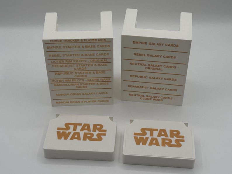 K&ouml;nnte beinhalten: Vier wei&szlig;e Star Wars Kartenboxen und zwei Kartenstapel. Die Boxen haben eine U-f&ouml;rmige Oberseite und Text in Orange. Die Kartenstapel haben das Star Wars Logo in Orange. Die Boxen listen Kartentypen wie "Empire Galaxy Cards" und "Rebel Starter & Base Cards".