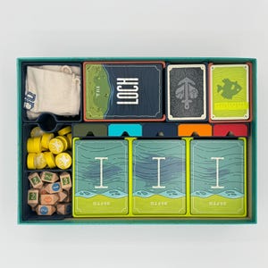 May include: A board game box interior containing a white cloth bag, a deck of cards labelled The Loch, a deck with an anchor illustration, a deck with a fish icon, three stacks of Depth cards, yellow circular tokens, and wooden number blocks.