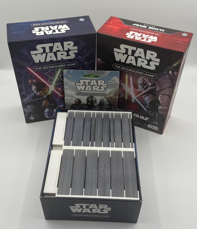 K&ouml;nnte beinhalten: Eine Sammlung von Star Wars Deckbuilding-Spielboxen und -Karten. Die Boxen sind marineblau und rot, mit dem Star Wars-Logo und Charakterillustrationen. Die Spielkarten sind grau und in einer schwarzen Box angeordnet.