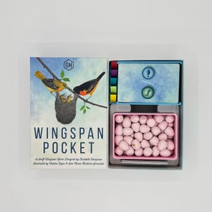 May include: Wingspan Pocket board game box featuring an illustration of two birds at a nest. The open box contains a blue card tray with circular fish and worm tokens, and a pink rectangular tray filled with small, speckled light pink eggs.
