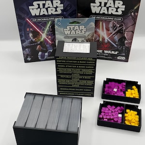 K&ouml;nnte beinhalten: Zwei Star Wars Deckbuilding-Spielboxen, eine kleine Kartenpackung und ein schwarzer Kartenhalter mit Karten werden gezeigt. Zwei schwarze Tabletts enthalten lila und gelbe Spielsteine. Die Boxen zeigen Bilder von Charakteren mit Lichtschwertern.