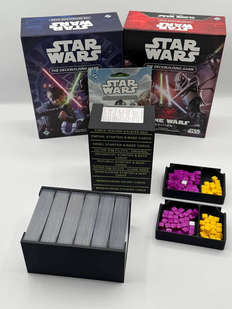 K&ouml;nnte beinhalten: Zwei Star Wars Deckbuilding-Spielboxen, eine kleine Kartenpackung und ein schwarzer Kartenhalter mit Karten werden gezeigt. Zwei schwarze Tabletts enthalten lila und gelbe Spielsteine. Die Boxen zeigen Bilder von Charakteren mit Lichtschwertern.