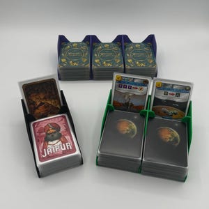 May include: A collection of board game card holders in black, green, and purple. The holders are filled with various game cards, including those from Jaipur and Wingspan. The cards feature illustrations and text.