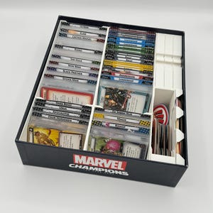 May include: A black Marvel Champions The Card Game box containing white plastic dividers organizing numerous playing cards. The cards are labeled with names like Ultron, Klaw, Captain Marvel, Rhino, She-Hulk, Iron Man, Black Panther, and Spider-Man.