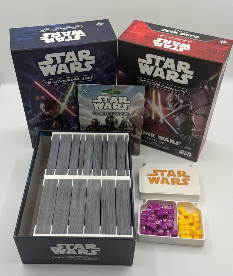 K&ouml;nnte beinhalten: Zwei Star Wars Deckbuilding-Spielboxen, eine blaue und eine rote, mit dem Spieltitel deutlich sichtbar. Die offene Box zeigt einen Kartenorganisator mit grauen Karten sowie Spielsteinen.