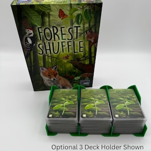 K&ouml;nnte beinhalten: Ein Brettspielkarton mit dem Titel "Forest Shuffle" und Illustrationen von Waldtieren. Drei Kartenstapel werden in einem gr&uuml;nen Halter gehalten. Der Text "Optional 3 Deck Holder Shown" befindet sich unten.