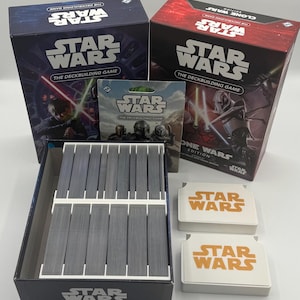 K&ouml;nnte beinhalten: Eine Sammlung von Star Wars Brettspielen. Das Bild zeigt zwei Spielboxen, eine Kartenpackung, eine Kartenbox und zwei wei&szlig;e Spielsteine mit dem Star Wars-Logo. Die Boxen sind blau und rot.