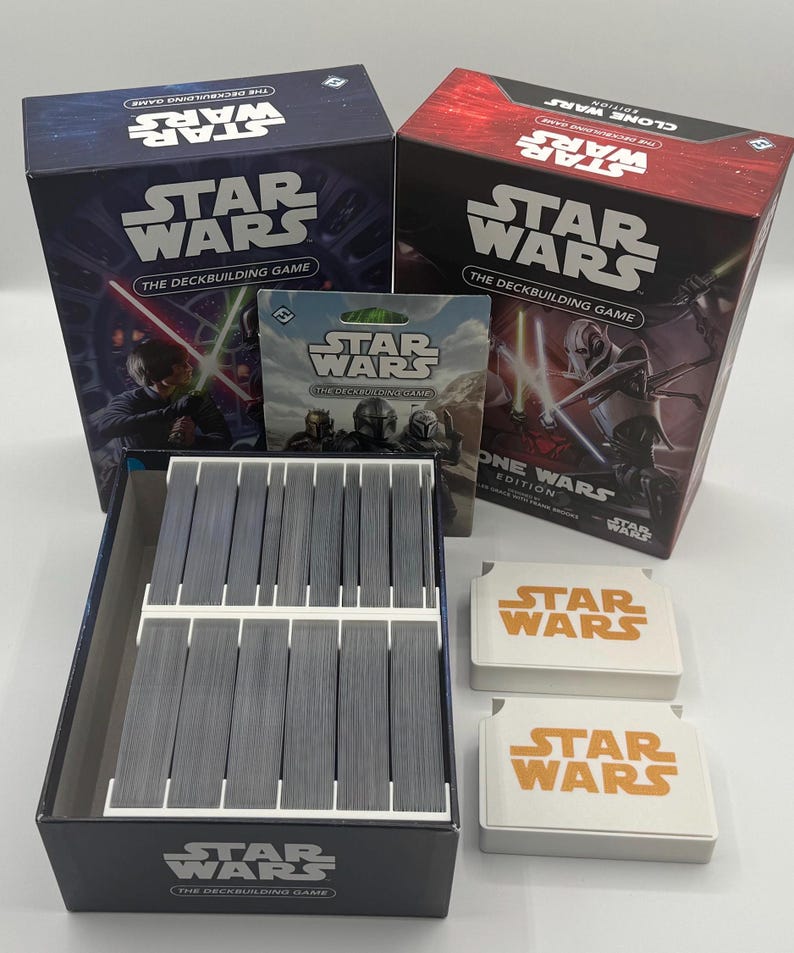 K&ouml;nnte beinhalten: Eine Sammlung von Star Wars Brettspielen. Das Bild zeigt zwei Spielboxen, eine Kartenpackung, eine Kartenbox und zwei wei&szlig;e Spielsteine mit dem Star Wars-Logo. Die Boxen sind blau und rot.