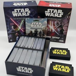 K&ouml;nnte beinhalten: Eine Sammlung von Star Wars Brettspielen. Das Bild zeigt zwei Spielboxen, eine Packung Karten, eine Kartenbox und zwei schwarze Kartenhalter. Die Boxen und Kartenhalter haben das Star Wars Logo in Wei&szlig; und Gelb.