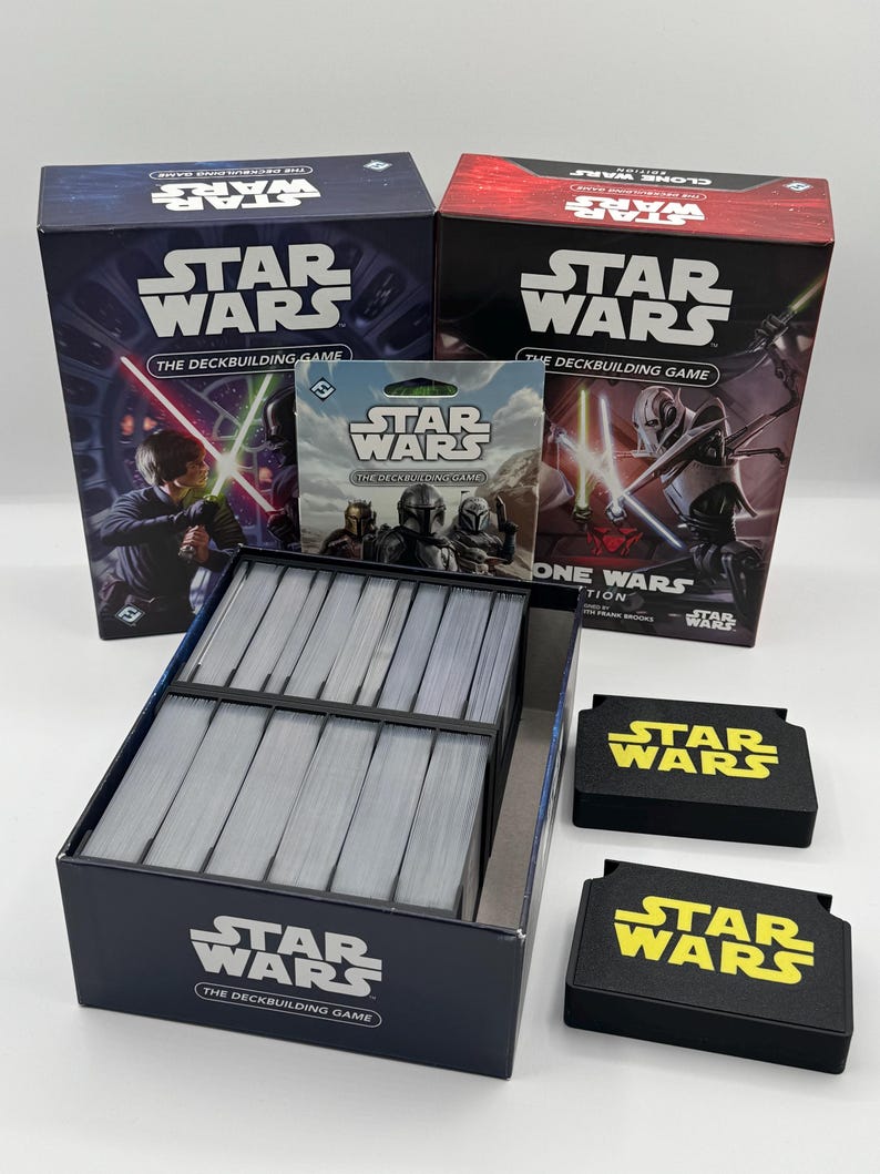 K&ouml;nnte beinhalten: Eine Sammlung von Star Wars Brettspielen. Das Bild zeigt zwei Spielboxen, eine Packung Karten, eine Kartenbox und zwei schwarze Kartenhalter. Die Boxen und Kartenhalter haben das Star Wars Logo in Wei&szlig; und Gelb.