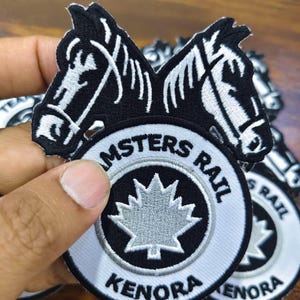 Op de afbeelding: Geborduurde patch met twee paardenkoppen boven een cirkelvormig ontwerp. Het ontwerp bevat de tekst "AMSTERS RAIL" en "KENORA" rond een grijs esdoornblad op een zwart-witte achtergrond.