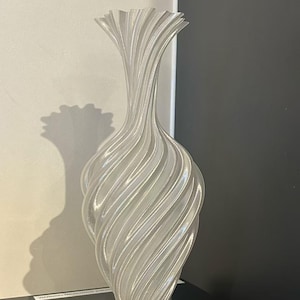 Könnte beinhalten: Eine hohe, klare Vase mit einem wirbelnden, gerippten Design. Die Vase hat einen ausgestellten oberen Teil und einen abgerundeten Boden. Das Licht fängt die Rippen ein und erzeugt Glanzlichter und Schatten. Die Vase besteht aus einem durchscheinenden Material.