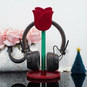 Può includere: Cuffie appoggiate su un supporto per cuffie rosso e verde a forma di rosa. Il supporto ha una base rossa e uno stelo verde che sostiene una rosa di velluto rosso. Le cuffie sono nere con dettagli marroni.
