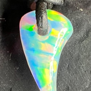 May include: A pear-shaped opal pendant with a rainbow iridescence, suspended from a knotted black leather cord. The gemstone displays a vibrant play of colors, including green, yellow, and blue. The pendant is a unique piece of jewelry.
