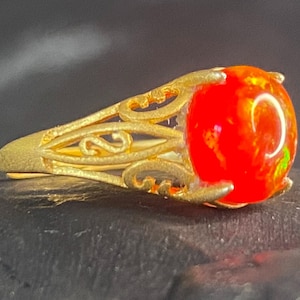 Pode incluir: Um anel dourado com uma pedra preciosa de opala vermelha oval e flamejante. O anel tem um design ornamental de filigrana na banda. A opala exibe cores vibrantes, com toques de verde e amarelo. O anel está sobre um fundo escuro.