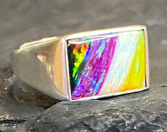 May include: A silver ring with a rectangular, iridescent stone. The stone displays a vibrant array of colors, including yellow, purple, and green, creating a rainbow effect. The ring has a simple, modern design.