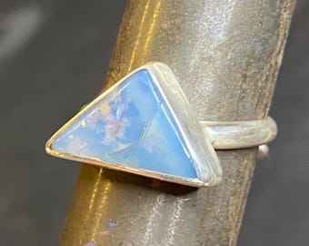 May include: A silver ring featuring a triangular, light blue opal gemstone. The opal has a milky appearance with subtle color variations. The ring is set in a simple, polished silver band.