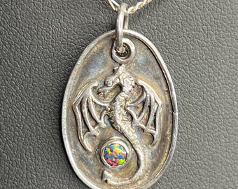 May include: A silver pendant necklace featuring an oval-shaped pendant with a detailed dragon design. The dragon has outstretched wings and a colorful, round stone at its center. The necklace chain is also silver.