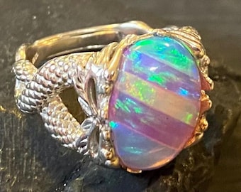 May include: A silver ring with an oval opal gemstone. The opal displays a vibrant play of color, with bands of green, purple, and white. The ring's setting has a detailed, textured design, possibly resembling a dragon or serpent.