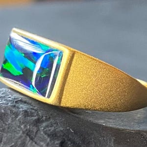 May include: A gold ring with a rectangular opal stone. The opal displays vibrant blue, green, and black colors. The ring has a textured band and a simple, modern design.