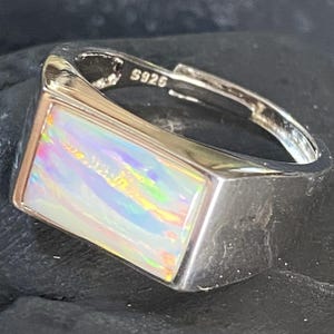 Peut inclure: Une bague en argent avec une pierre d'opale rectangulaire. L'opale présente un jeu de couleurs vives, notamment des teintes de rose, de bleu et de jaune. La bague est estampillée "S925".