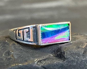 May include: A silver ring with a rectangular, iridescent stone. The stone displays hues of green, blue, and purple. The ring features a Greek key design on the sides, adding a touch of classic style to the jewelry.