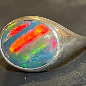 May include: A silver ring featuring a large, oval opal. The opal showcases vivid colours: red, orange, yellow, green, and blue. The ring's band is smooth and polished, providing a striking contrast with the colourful gemstone.