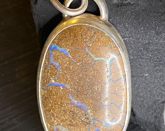 May include: Oval pendant with a silver-colored bail. The pendant features a brown stone with blue and green veins. The stone is set in a silver-colored bezel. The pendant is likely a piece of jewelry.