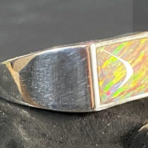 May include: A silver ring with a rectangular, iridescent opal stone. The opal displays a mix of colors including yellow, green, and red. The ring has a polished, brushed metal finish.