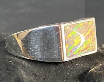 May include: A silver ring with a rectangular, iridescent opal stone. The opal displays a mix of colors including yellow, green, and red. The ring has a polished, brushed metal finish.