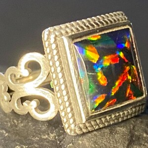 Könnte beinhalten: Ein silberner Ring mit einem quadratischen schwarzen Opal-Edelstein, der in einer silbernen Fassung sitzt. Der Ring hat ein dekoratives Band mit einem Schriftrollenmuster.