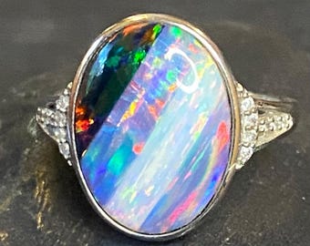 May include: Oval-shaped opal ring with a silver band. The opal displays a vibrant play of color, including blues, greens, and reds. Small, clear stones are set on either side of the opal, adding sparkle to the ring.