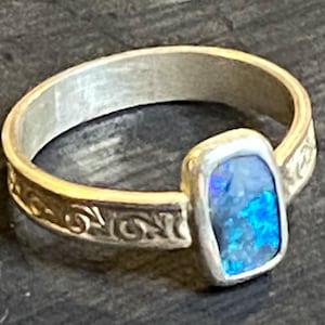 May include: A silver ring with an opal stone. The opal is a rectangular shape with shades of blue and purple. The band has a decorative pattern on the sides.
