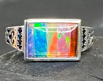 May include: A silver ring with a rectangular, multi-colored opal stone. The opal displays a vibrant spectrum of colors, including blue, green, orange, and red. The ring has intricate filigree detailing on the sides and small black stones.