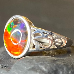 May include: A silver ring featuring an oval opal with a spectrum of colours, including orange, green, and purple. The ring's band has an openwork pattern. The ring is set against a dark backdrop.