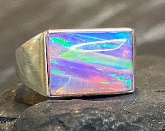May include: A silver ring with a rectangular opal stone. The opal displays a vibrant play of color, including blue, green, purple, and pink hues. The ring has a simple, modern design, with a polished silver band.