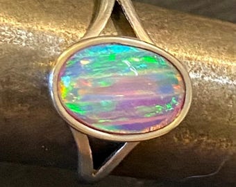 May include: A silver ring with an oval opal gemstone. The opal displays a vibrant play of color, including green, blue, and purple hues. The ring's band has a unique, open design, with a triangular shape on each side of the gemstone.