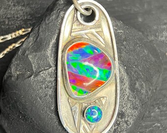 Può includere: Ciondolo in argento con un'opale multicolore vibrante. Il ciondolo ha una forma ovale con disegni incisi e una piccola pietra rotonda blu-verde nella parte inferiore. L'opale mostra uno spettro di colori.