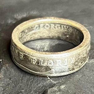 May include: A silver ring with engraved text, including "ONE FLORIDA" and "GEORGIVS". The ring has a worn, vintage appearance. The ring is a band style.