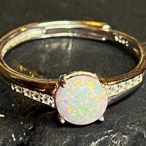 May include: A silver ring with a central, round opal gemstone. The opal has an iridescent quality, displaying colours like pink, green, and yellow. The band has small, clear accent stones. The ring has a polished finish.